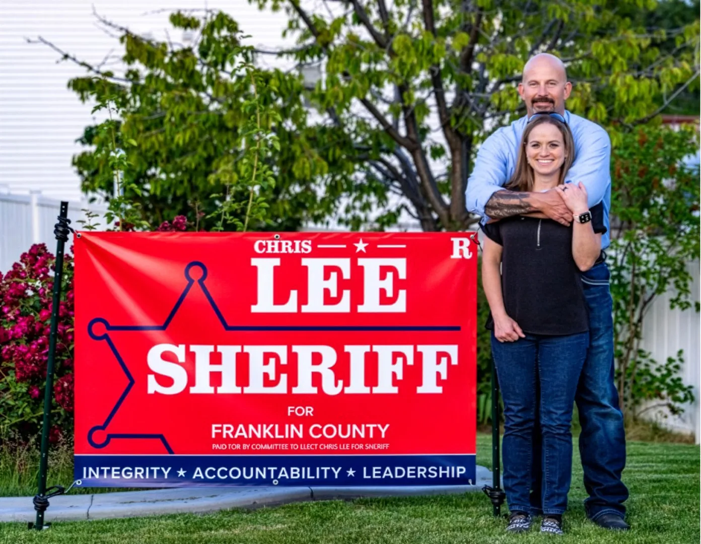 Chris Lee and his wife Julie standing beside a red Lee for Sheriff yard sign