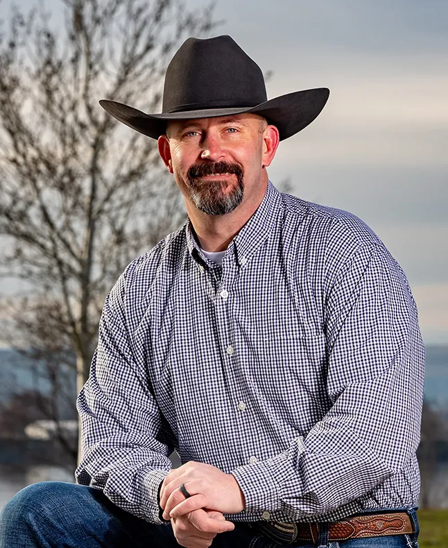 Chris Lee in a cowboy hat, flannel shirt and jeans, kneeling outdoors